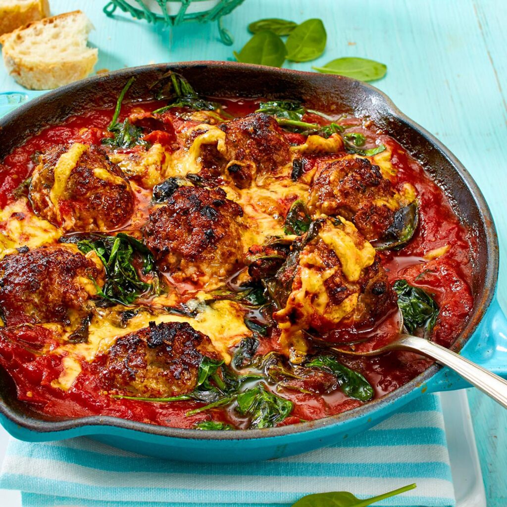 Spinach garlic meatballs baked in a rich tomato sauce with melted cheese, served hot in a skillet with fresh spinach leaves