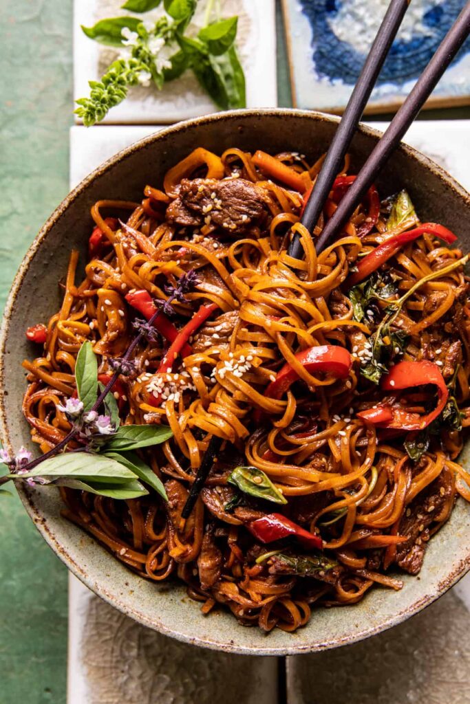 Korean beef noodles with tender beef slices, stir-fried noodles, red peppers, fresh herbs, and sesame seeds served in a bowl with chopsticks