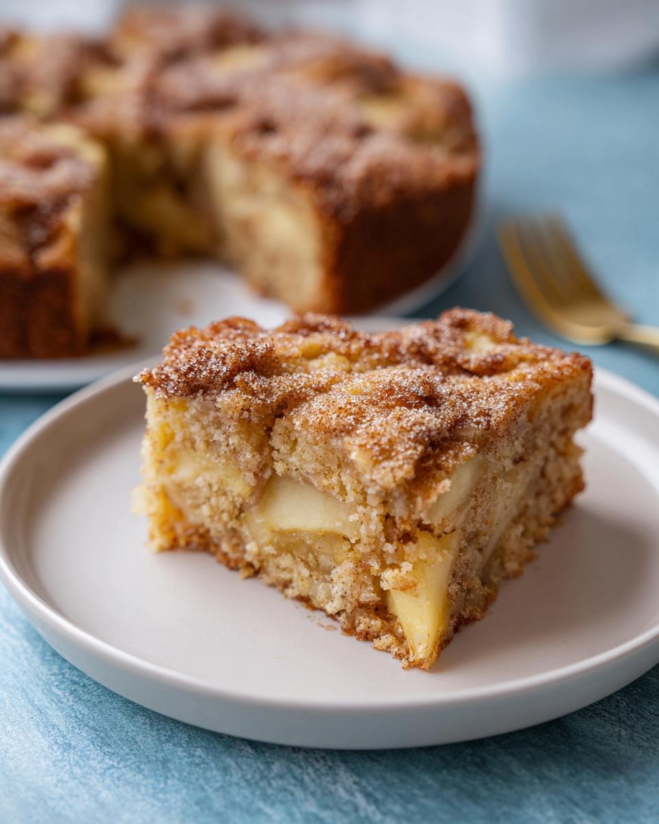 Ein Stück Apfelkuchen mit Zimt-Zucker-Topping auf einem weißen Teller, mit dem Restkuchen im Hintergrund.