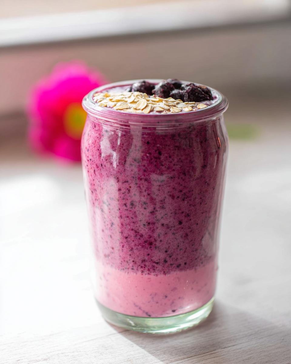 Ein hoher Glasbehälter gefüllt mit einem geschichteten Gesunder Frühstücks-Smoothie mit Joghurt, garniert mit Haferflocken und Beeren.